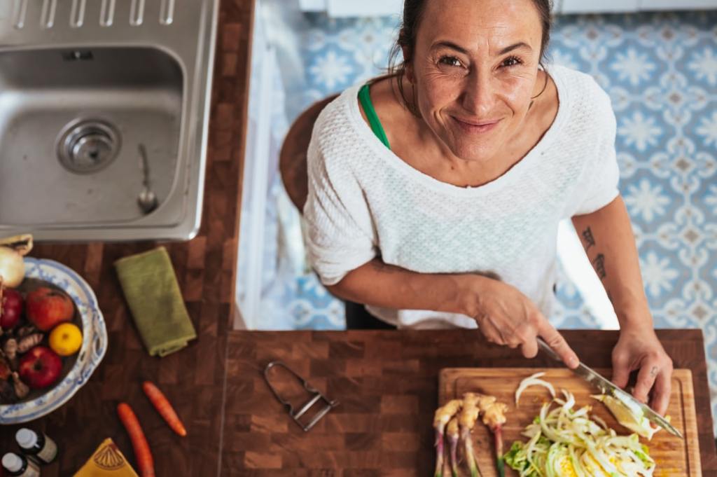 A smiling woman in a white top chopping vegetables at the kitchen table and enjoying the art of cooking